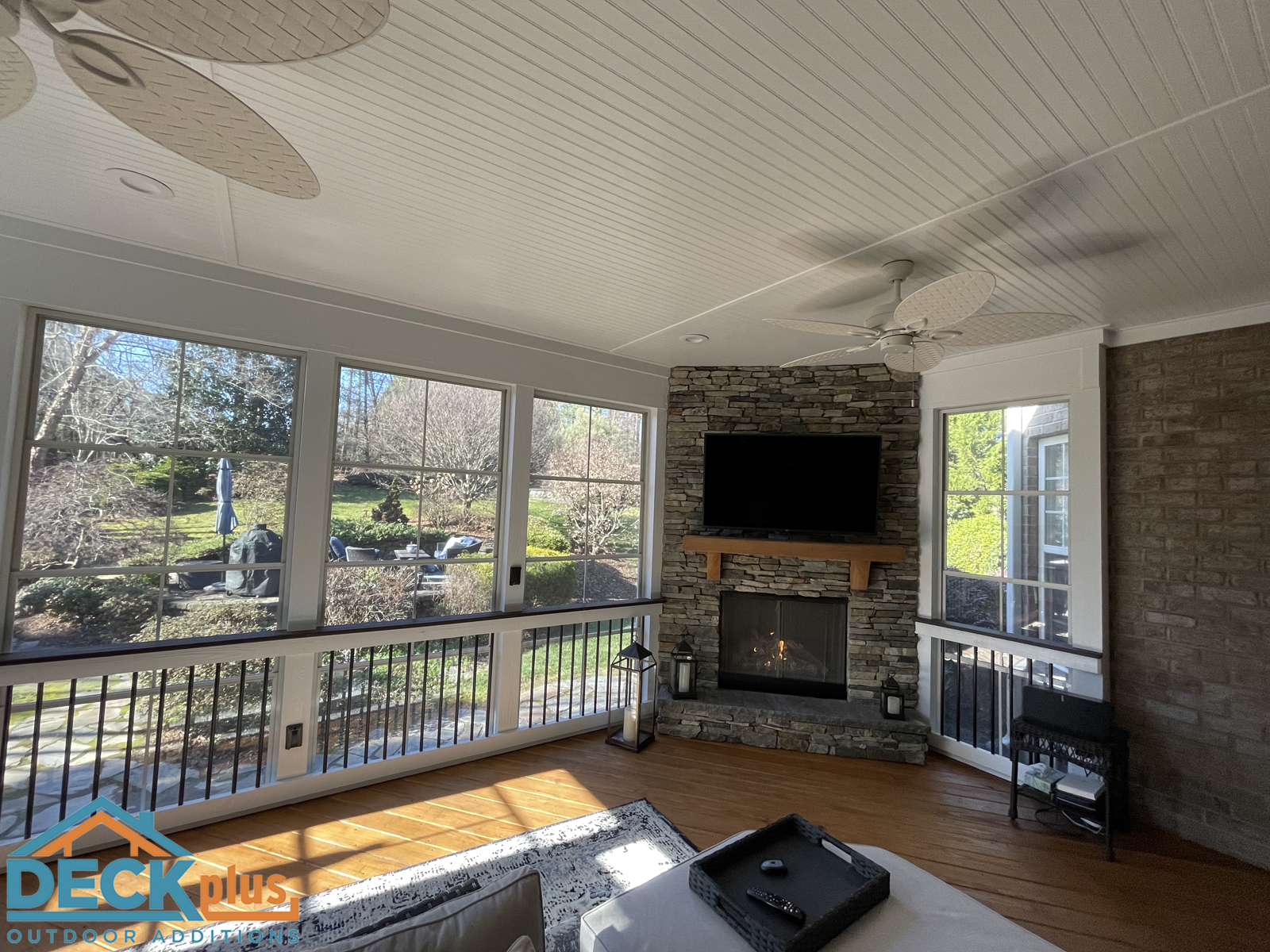 Screened-in-porch with Trex composite floors and EzeVue windows. Fireplace in corner with stone surround. Two white ceiling fans. View of trees outside.