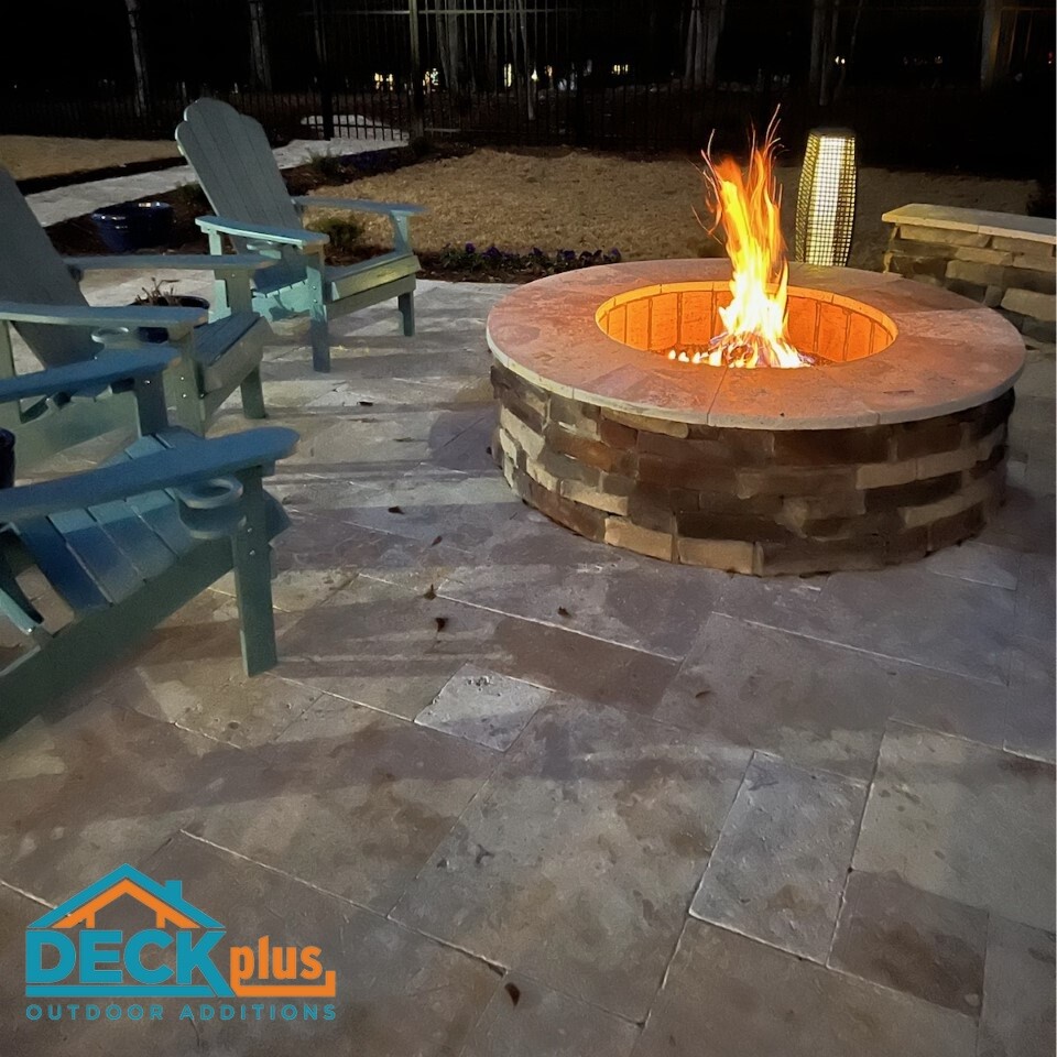 Patio and built-in stone fire pit in a backyard at night. Fire in the fire pit, with three blue Adirondack chairs around one side. Deck Plus logo in bottom left corner.