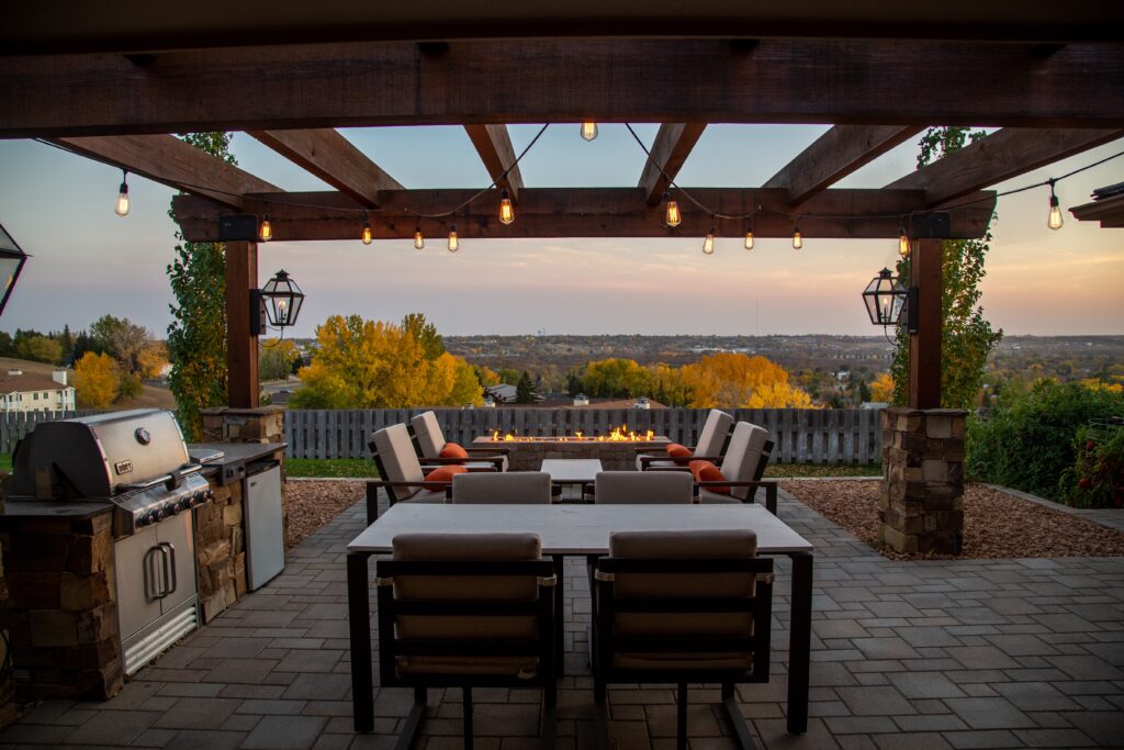 A large wooden pergola over outdoor kitchen in backyard with view overlooking a town at sunset.