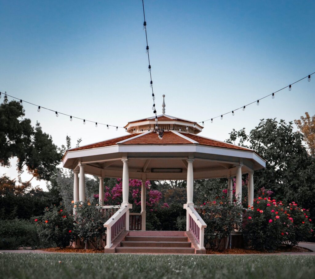 Gazebo on a manicured lawn, surrounded by flowers, and string lights attached and extending from the top of the roof. Trees and blue sky in background.